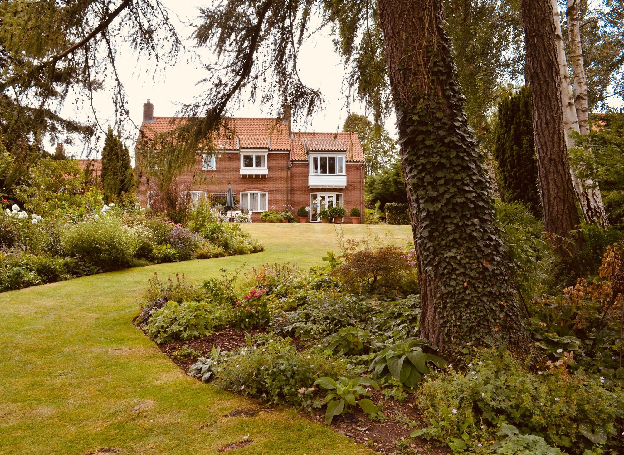 An English garden on a summer afternoon — mown lawn, deep planted borders and a mature cedar in front of a red-brick house