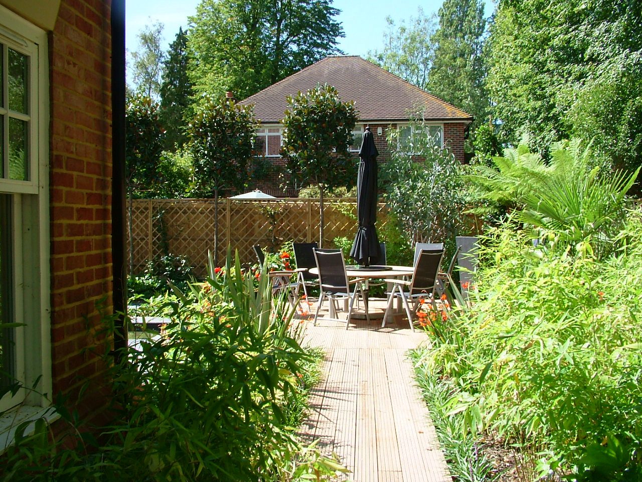 Timber decking wrapped around a brick house and softened with layered planting — table, chairs and parasol sitting level on boards laid to the fall of the garden.
