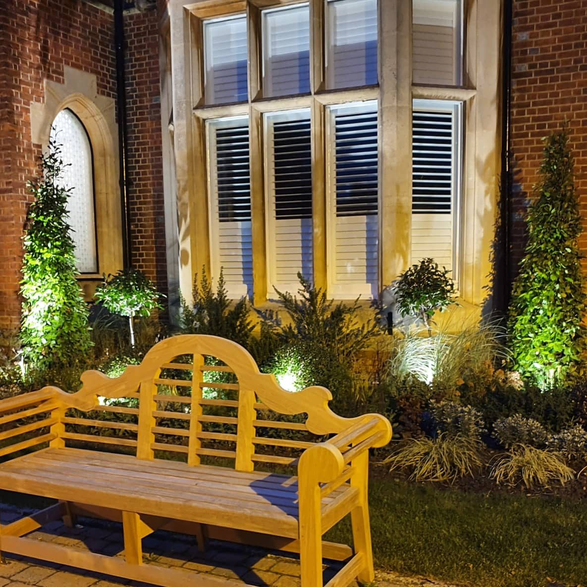 The same garden after dark: uplighting through clipped shrubs against a period brick and stone frontage, with a painted bench lit as the focal point.