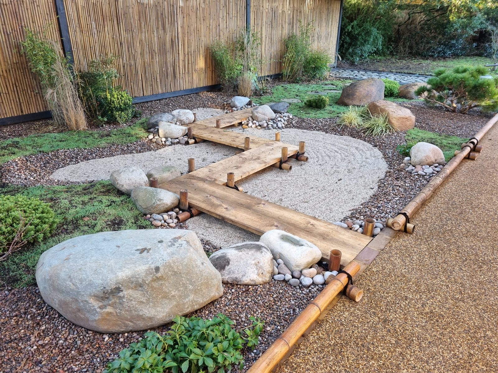 A gravel and boulder garden with railway-sleeper stepping treads, edged in timber against a bamboo screen — low-maintenance hard landscaping on a difficult, shady strip.