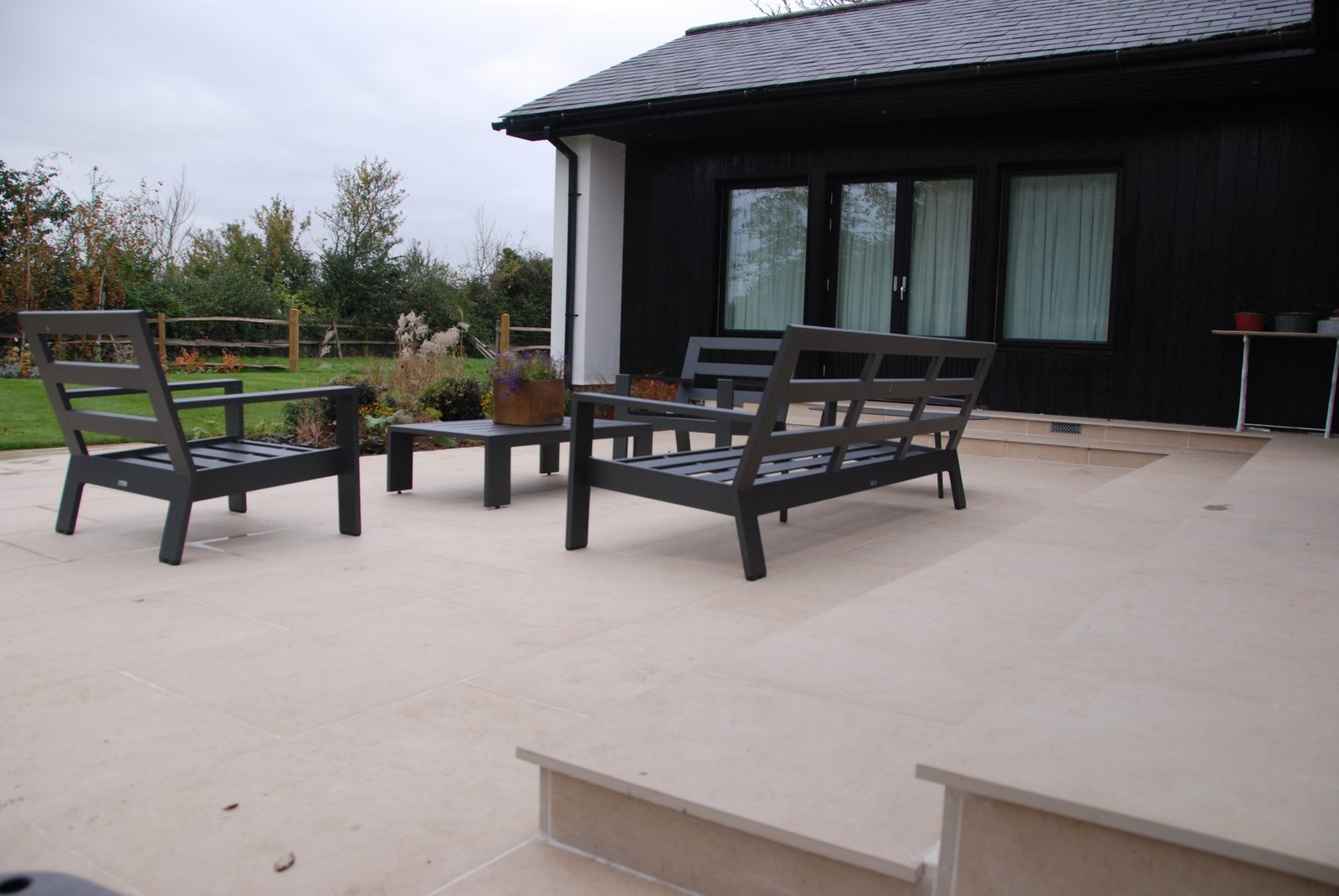 The same house from the garden: paving carried down in broad steps so the terrace meets the lawn without a lip, with furniture set out on the level.