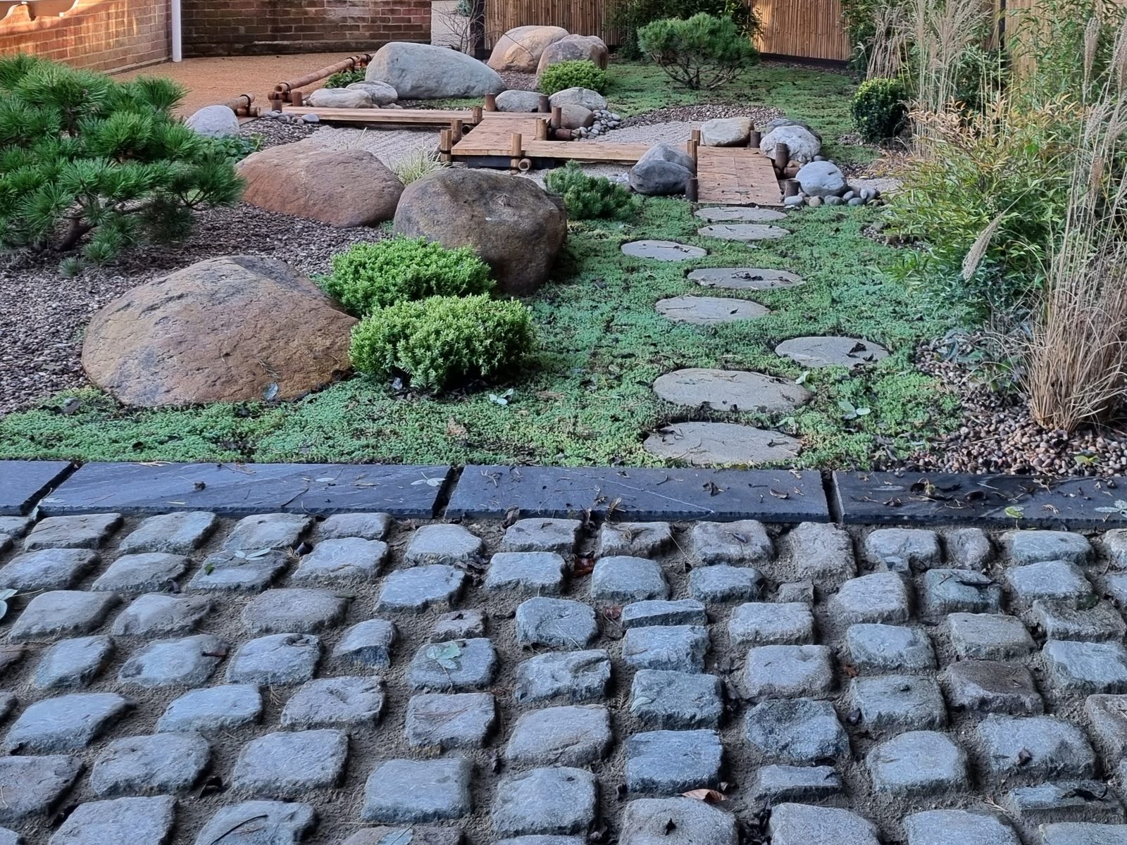 Stone setts running up to a stepping-stone path through a planted rockery, with boulders and low shrubs used to hold a bank and give it winter structure.